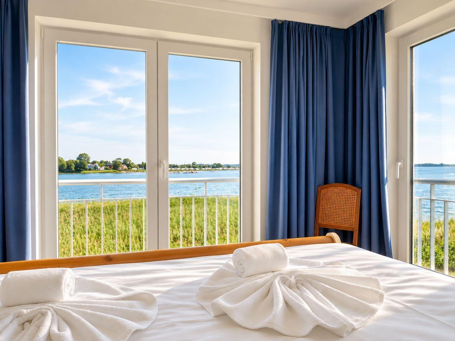 Schlafzimmer mit Blick auf Wasser und Grünflächen durch große Fenster.