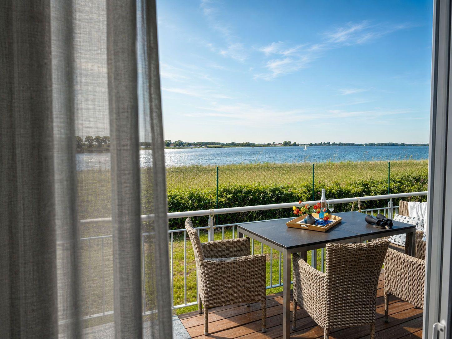 Terrasse mit Tisch und Stühlen bietet Blick auf See und Grünflächen.
