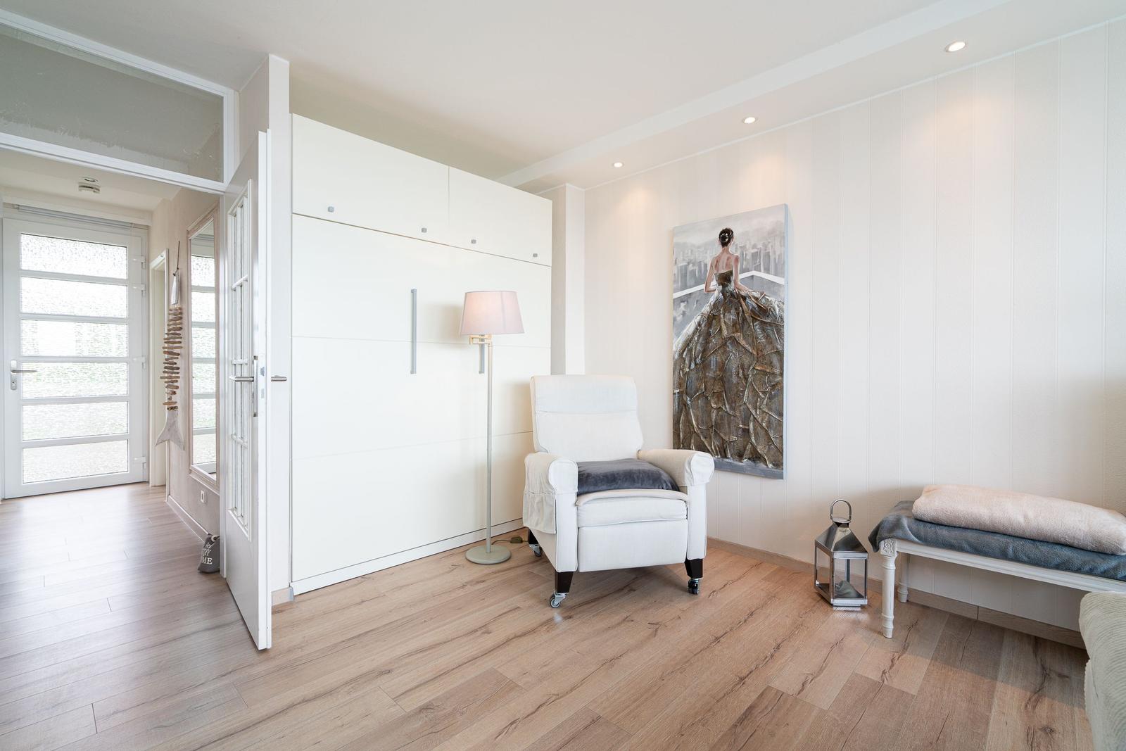 Ein Raum mit weißem Schrank, Sessel und Wandbild. Holzdielenboden und weiße Wände.