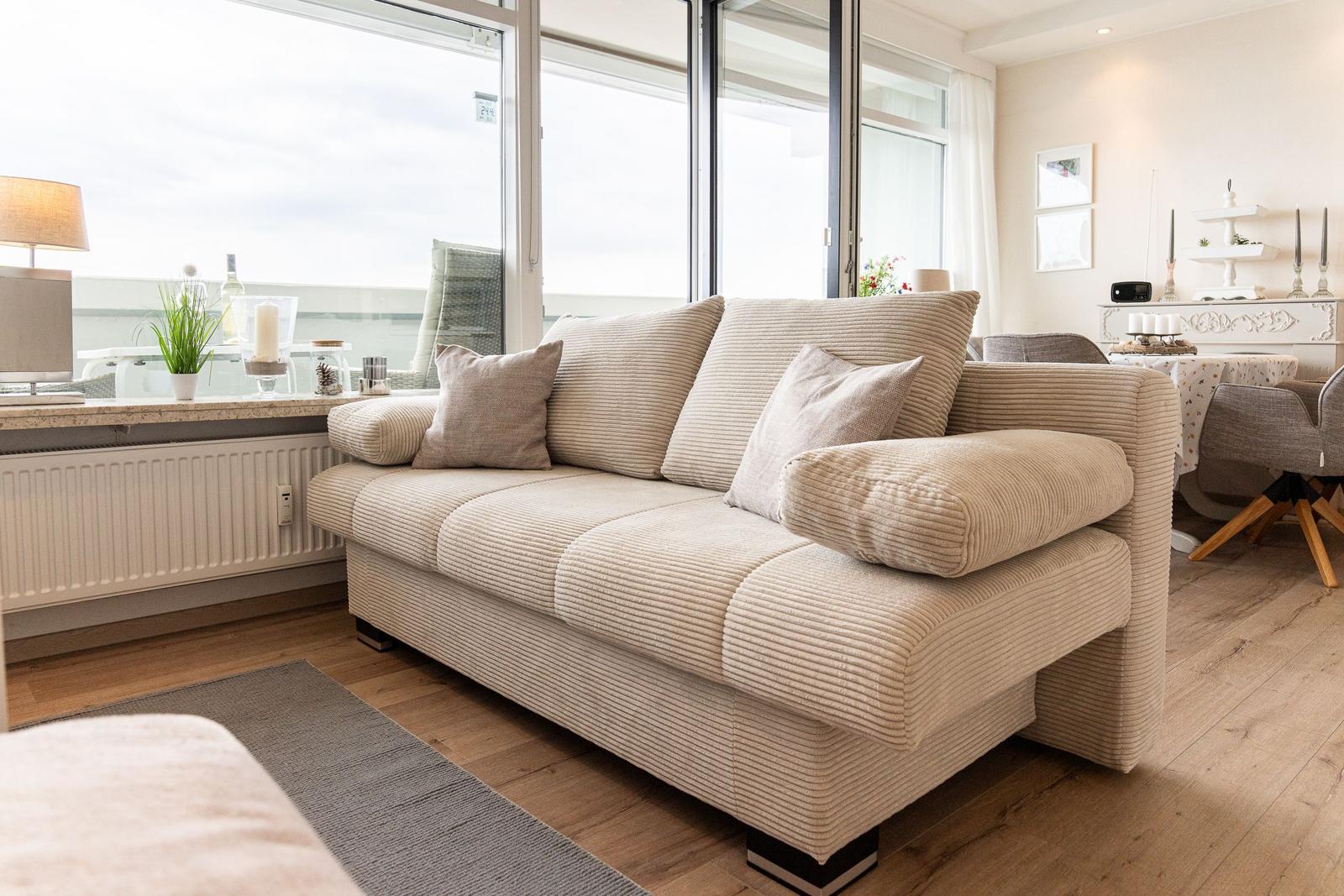 Ein Wohnzimmer mit einem beigen Sofa, Holzboden und großen Fenstern.