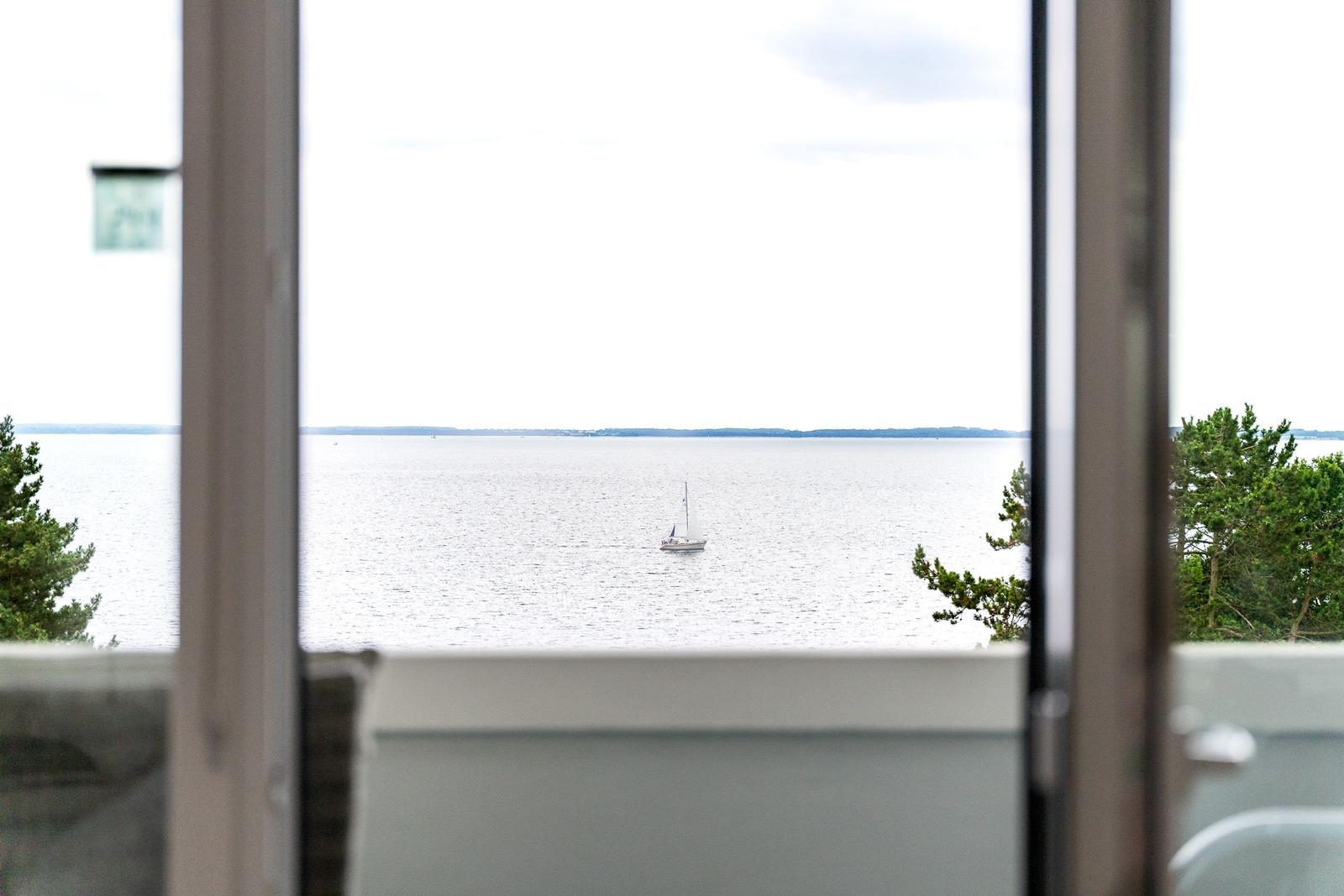 Vom Fenster aus Blick auf See mit Segelboot und Bäumen im Vordergrund.