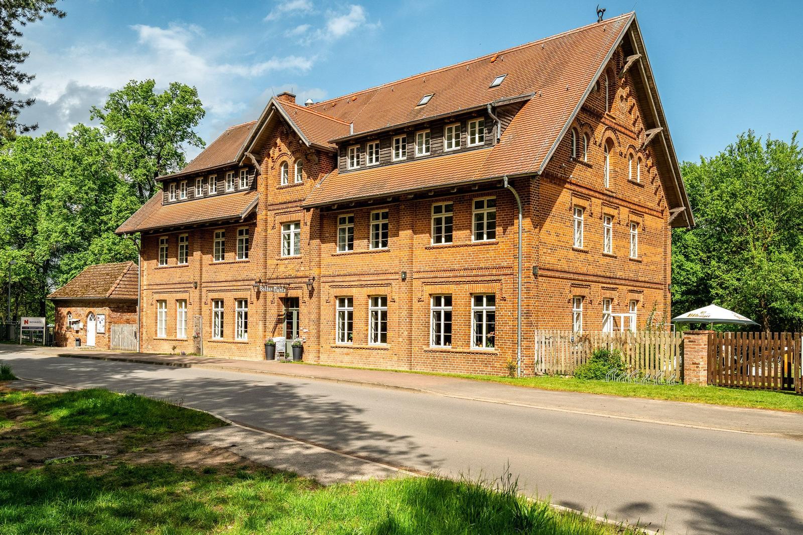 Rotes Backsteingebäude mit Dachgauben und Fenstern, umgeben von Bäumen und einem Zaun.