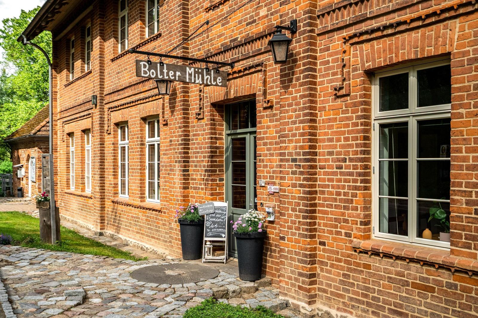 Roter Backsteinbau mit Eingangstür und Schild Bolter Mühle. Kiesweg und Blumenkübel.