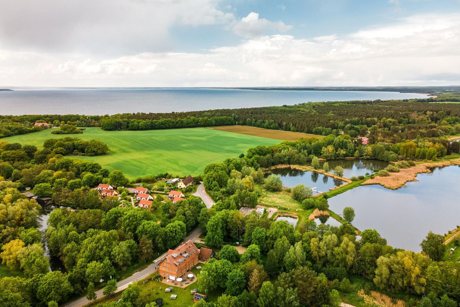 Landschaft mit See, Wiesen und Bäumen. Mehrere Häuser sind sichtbar.