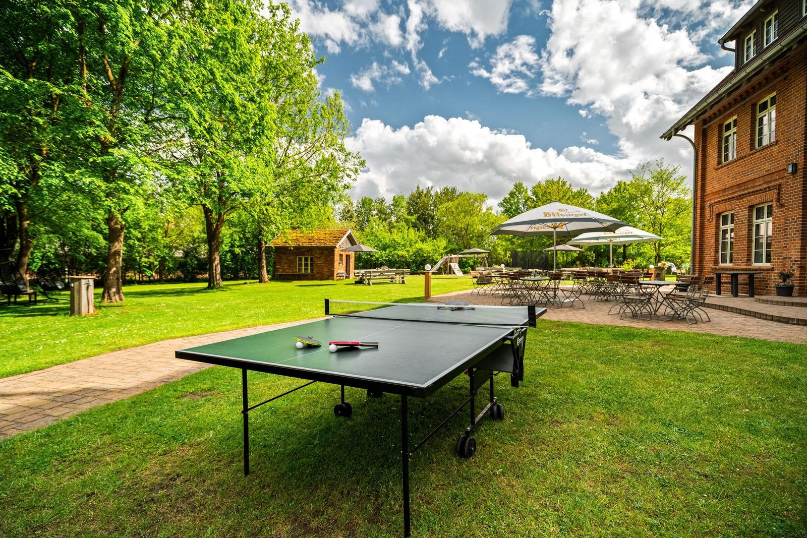 Tischtennisplatte auf Grünfläche, Terrasse mit Tischen und Sonnenschirmen, roter Backsteinbau im Hintergrund.