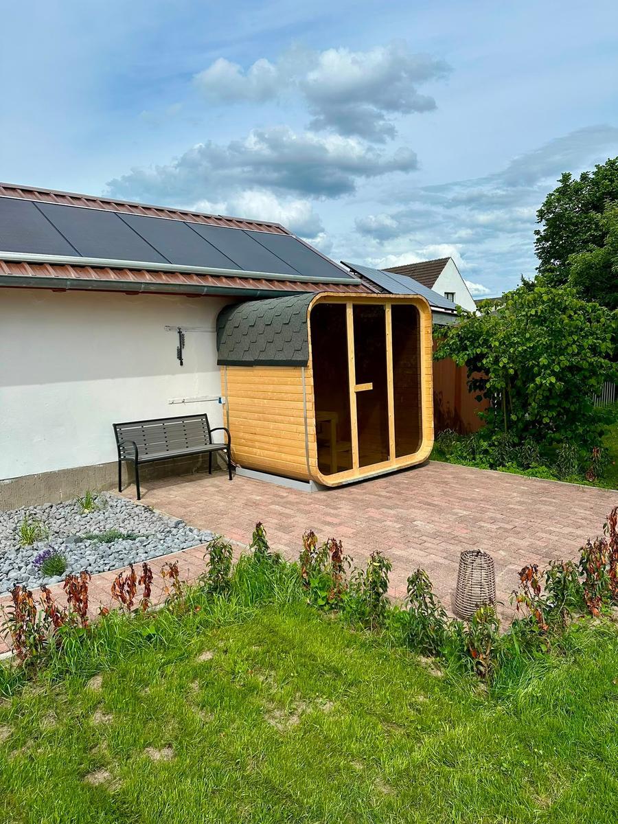 Holzsauna mit Glasfront auf Terrasse neben Haus mit Solaranlage.
