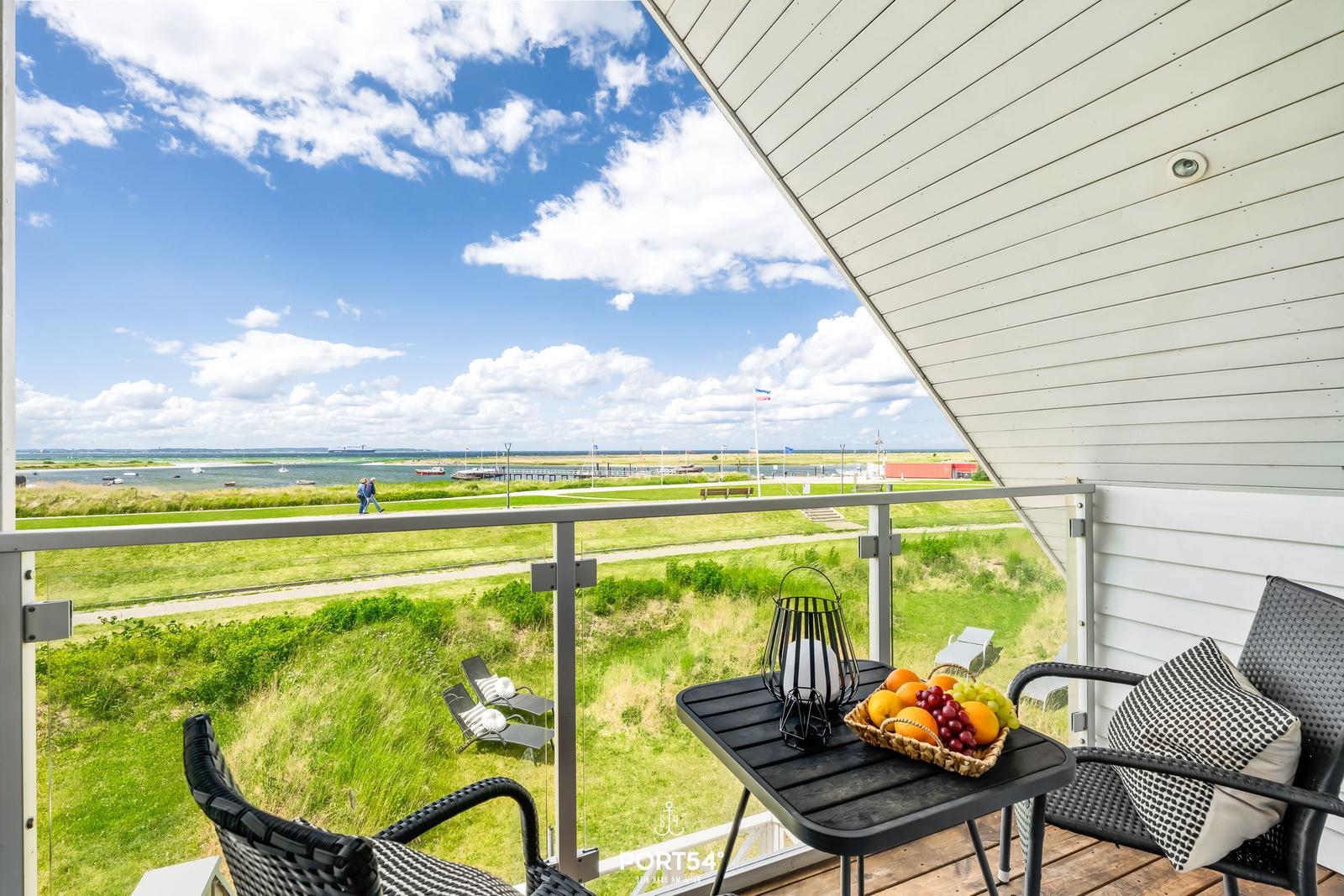 Terrasse mit Tisch, Stühlen und Obst. Blick auf Grünflächen und Wasser.