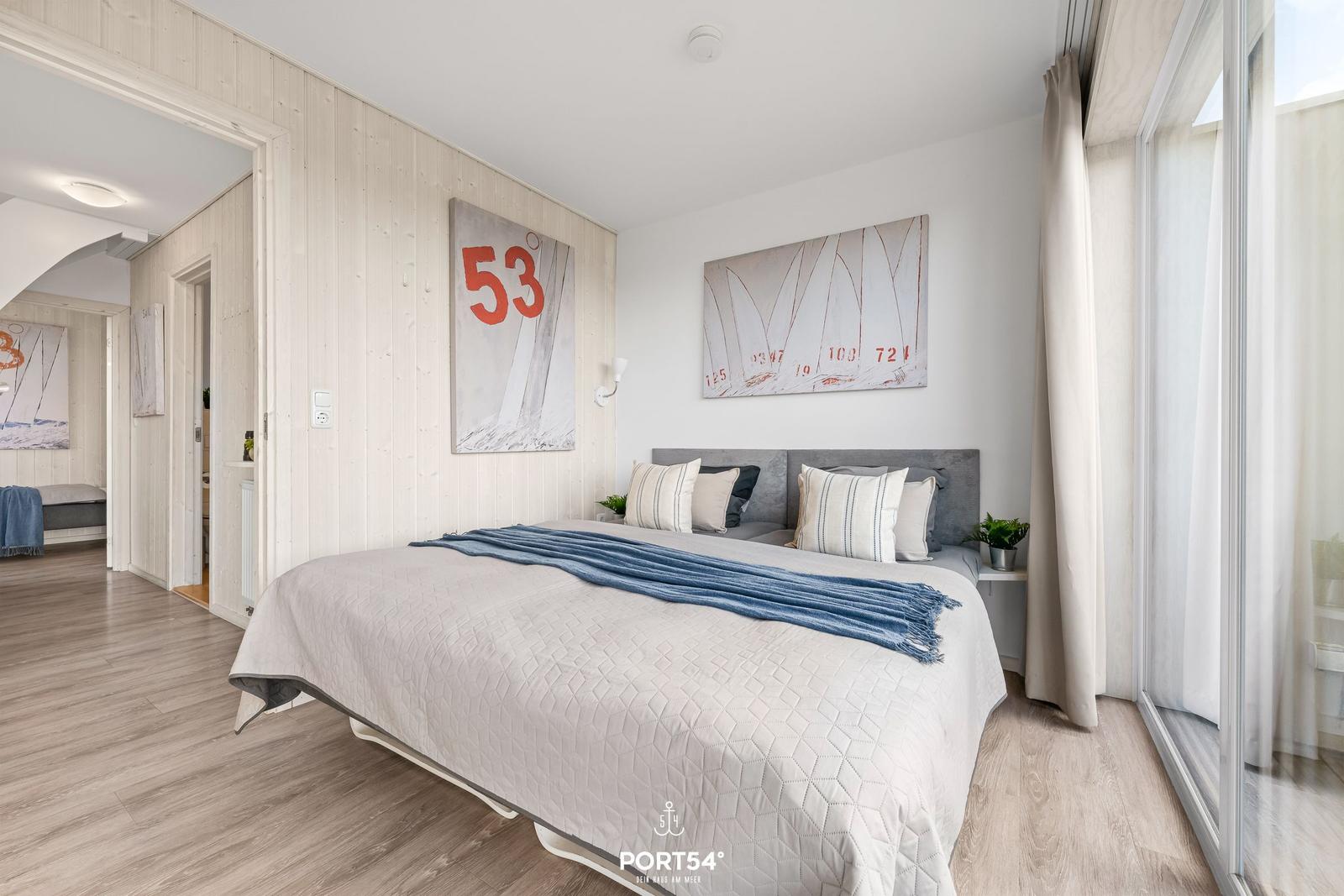 Schlafzimmer mit Bett, Wandbildern und großen Fenstern.