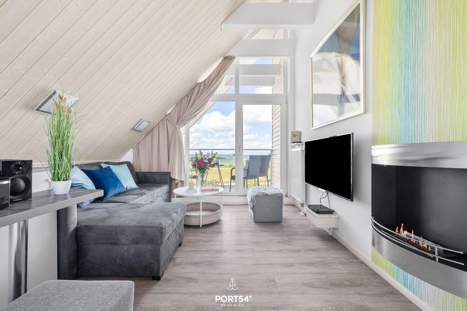 Wohnzimmer mit Sofa, Fernseher und Kamin. Blick auf Balkon mit Sicht auf Meer.