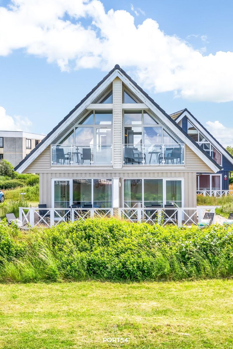 Modernes Haus mit großem Balkon und Glasfronten, umgeben von Grünflächen und einem klaren Himmel.