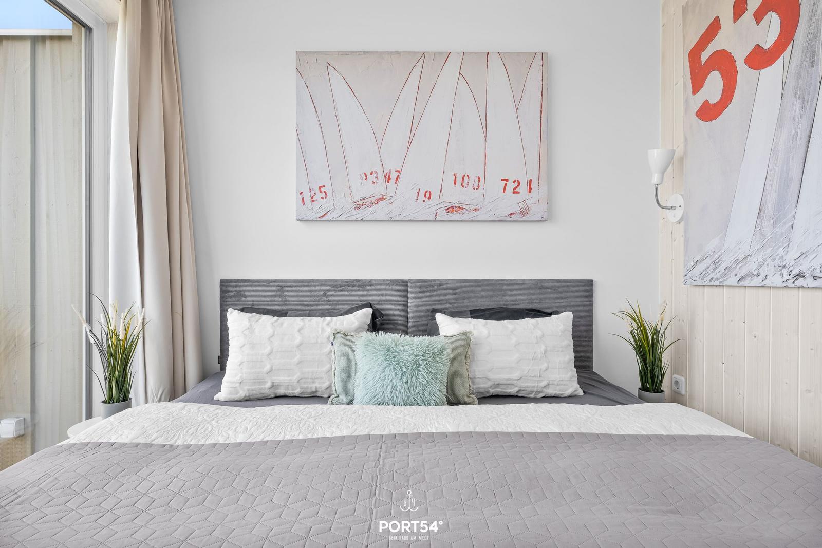 Schlafzimmer mit Bett, Wandbildern und Pflanzen. Fenster mit Vorhängen.