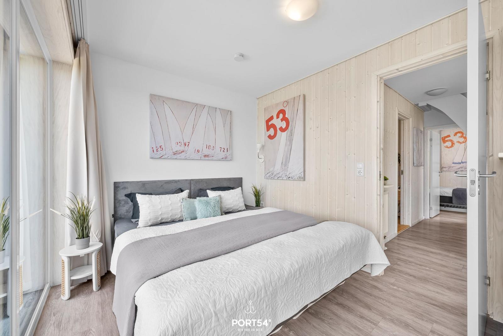 Schlafzimmer mit Bett, Wandbildern und Holzwand. Tür führt in angrenzenden Raum.
