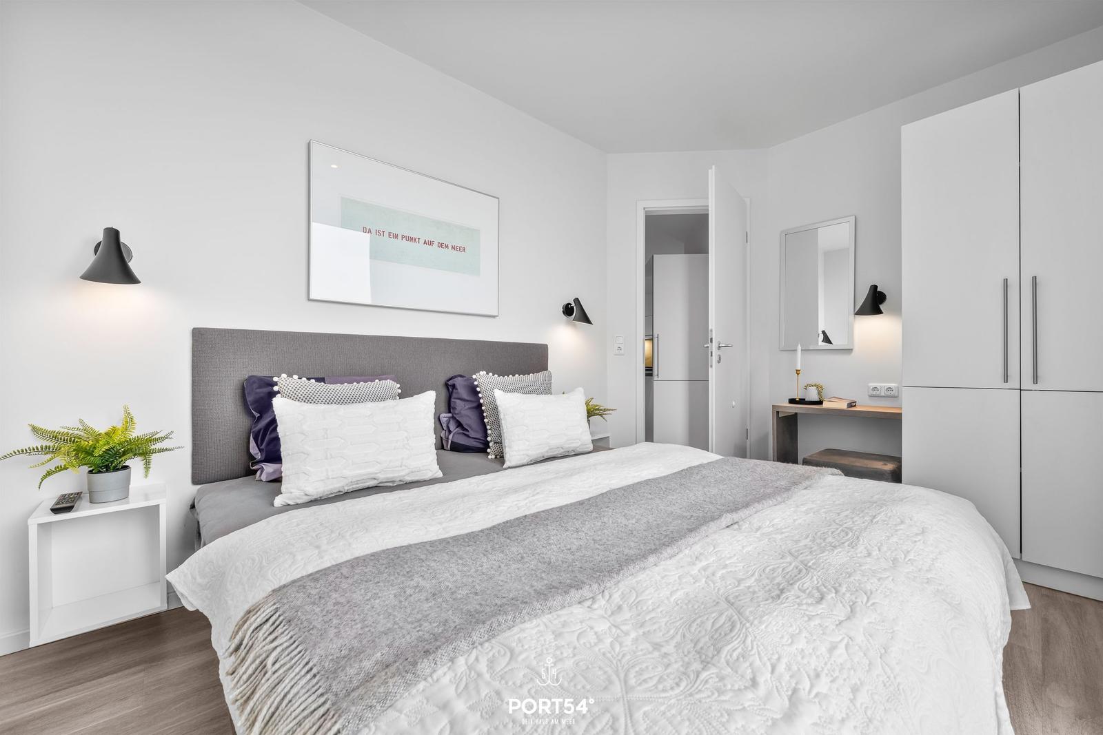 Schlafzimmer mit Bett, Kleiderschrank und Schreibtisch. Wandbild und Pflanzen sind sichtbar.