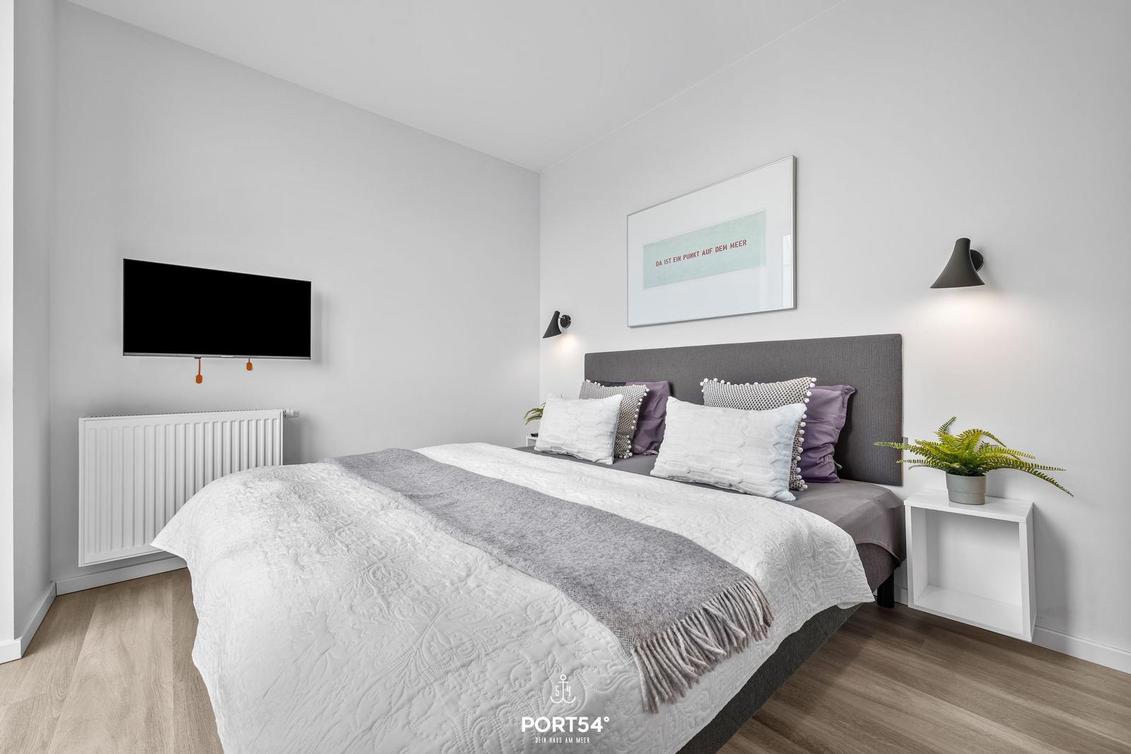 Schlafzimmer mit Bett, Fernseher und Wandlampe.