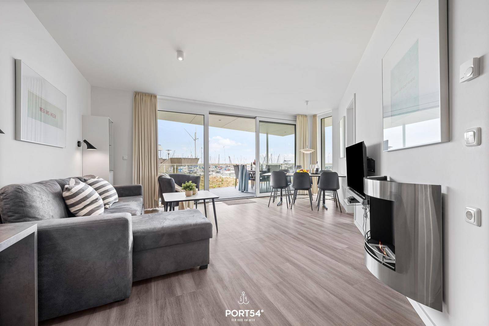 Wohnzimmer mit Sofa, Esstisch und Blick auf Hafen. Wandkamin und große Fenster.