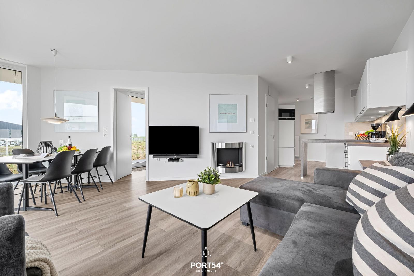 Wohnbereich mit Sofa, Fernseher und Kamin. Küche und Essbereich sind sichtbar.