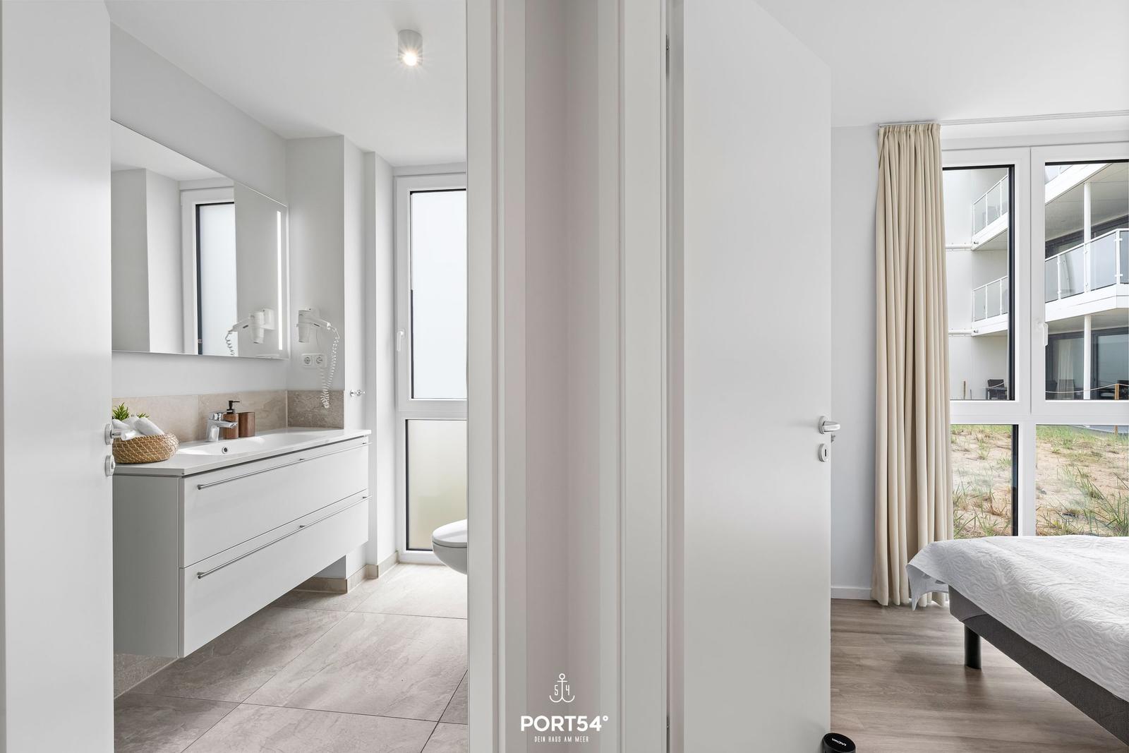 Badezimmer mit Waschbecken und Spiegel. Schlafzimmer mit Bett und Fenster.
