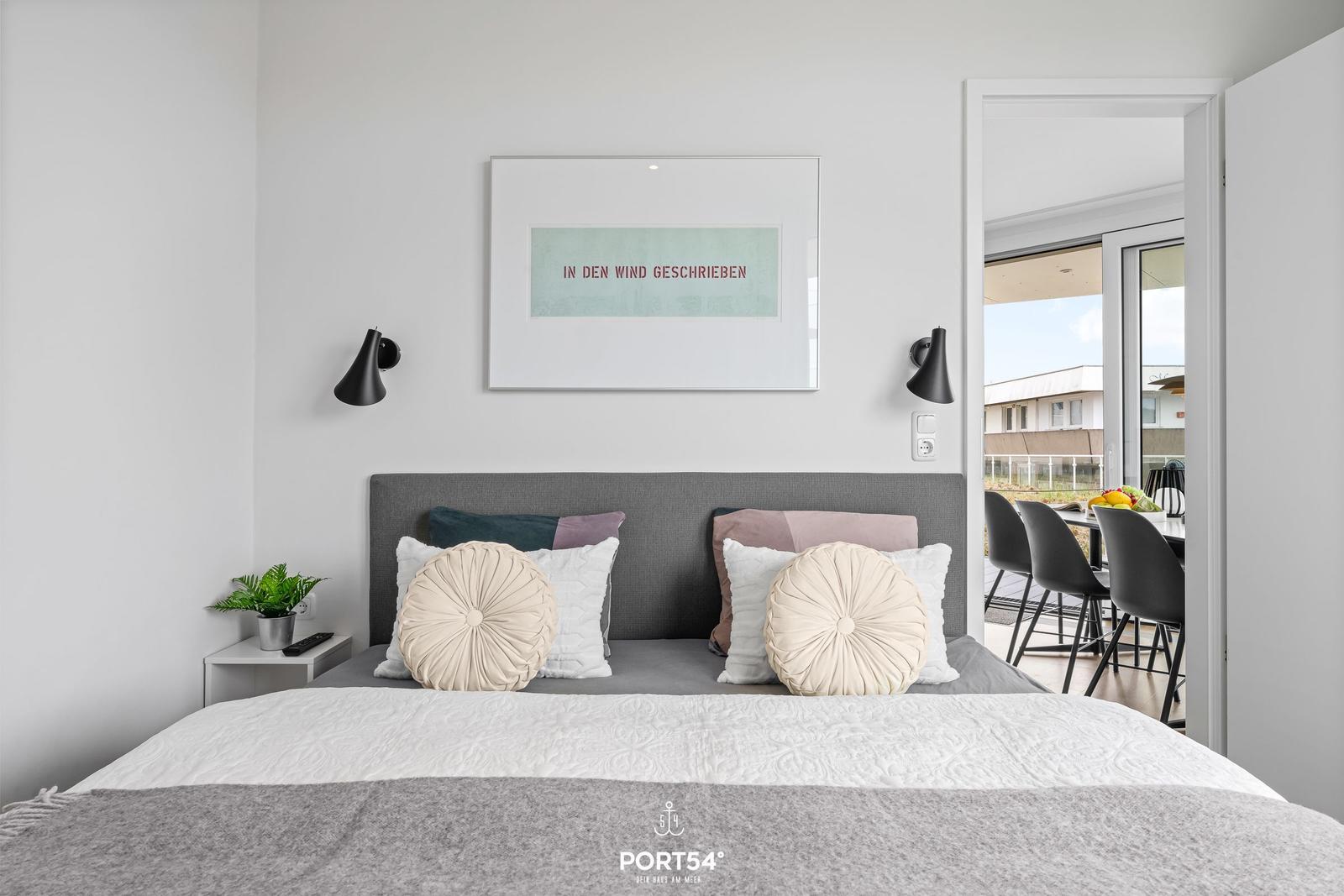 Schlafzimmer mit Bett, Kissen und Wandbild. Durchgang zur Terrasse mit Tisch und Stühlen.