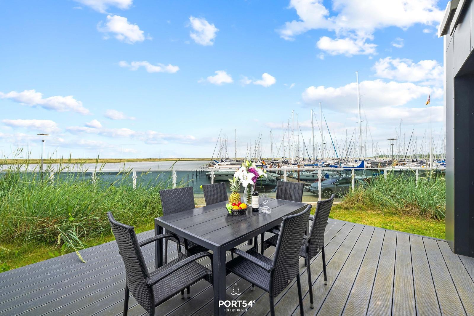 Terrasse mit Tisch und Stühlen, Blick auf Hafen mit Booten und Gras.