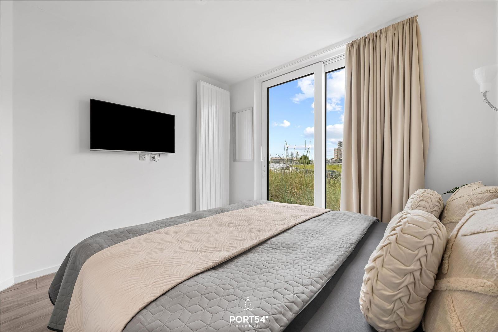 Schlafzimmer mit Bett, TV und großem Fenster mit Blick auf Grünfläche.