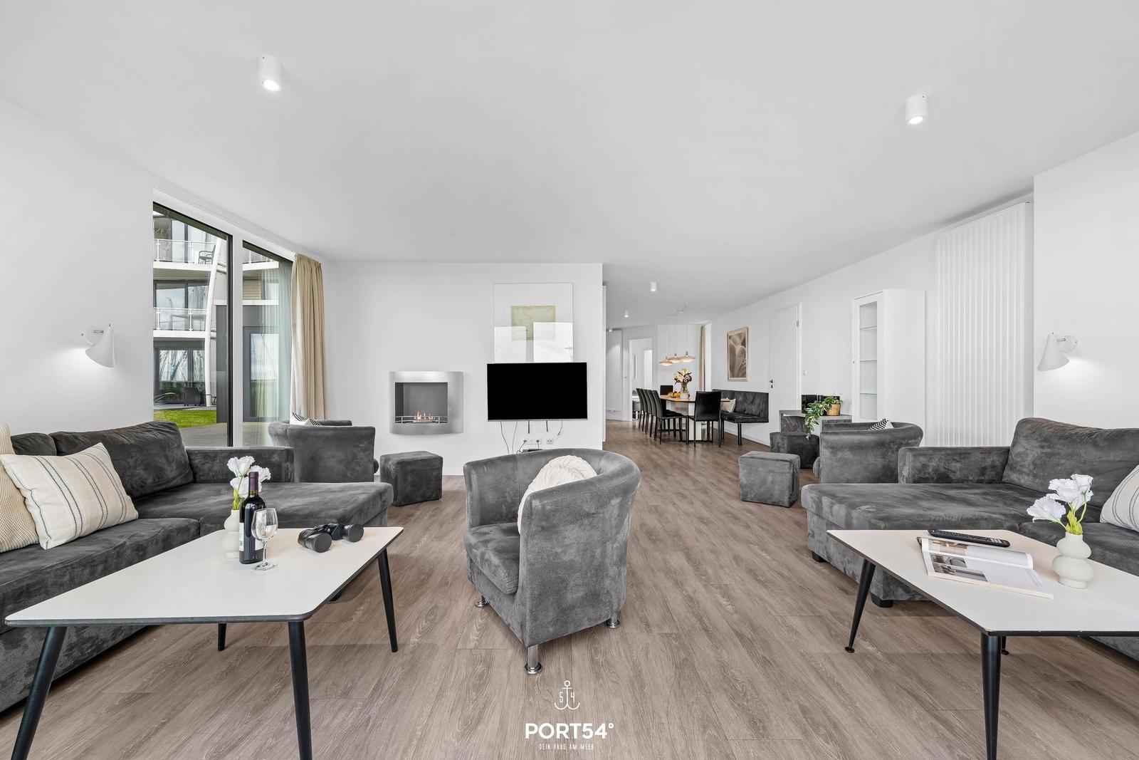 Wohnbereich mit grauen Sofas, Fernseher und Kamin. Holzboden und große Fenster.
