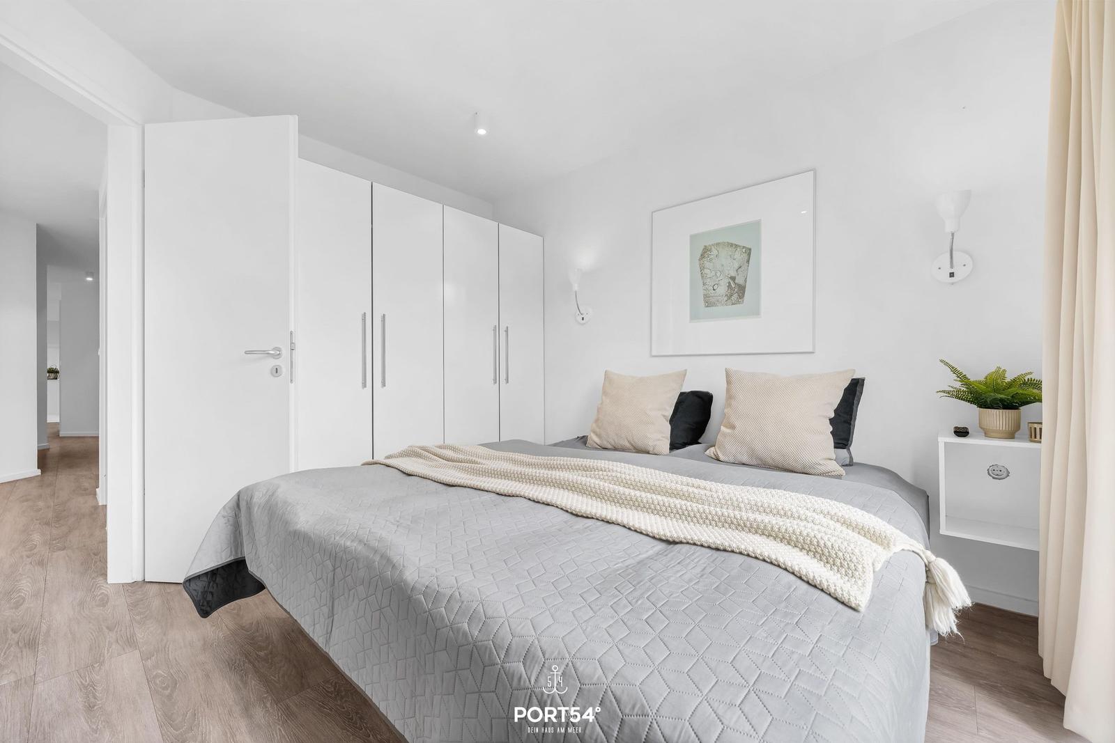 Schlafzimmer mit Bett, Kleiderschrank und Nachttisch. Wandbild und Pflanze sichtbar.