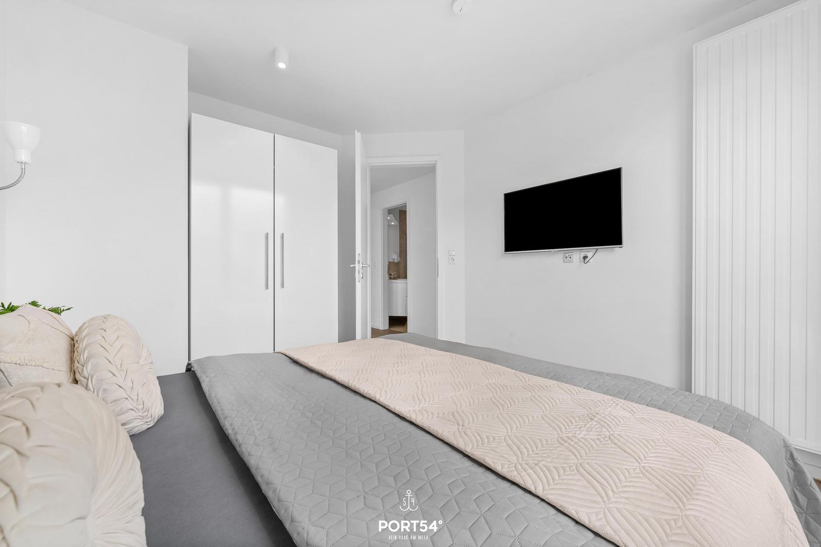 Schlafzimmer mit Bett, Kleiderschrank und Wandfernseher. Tür führt in Bad.
