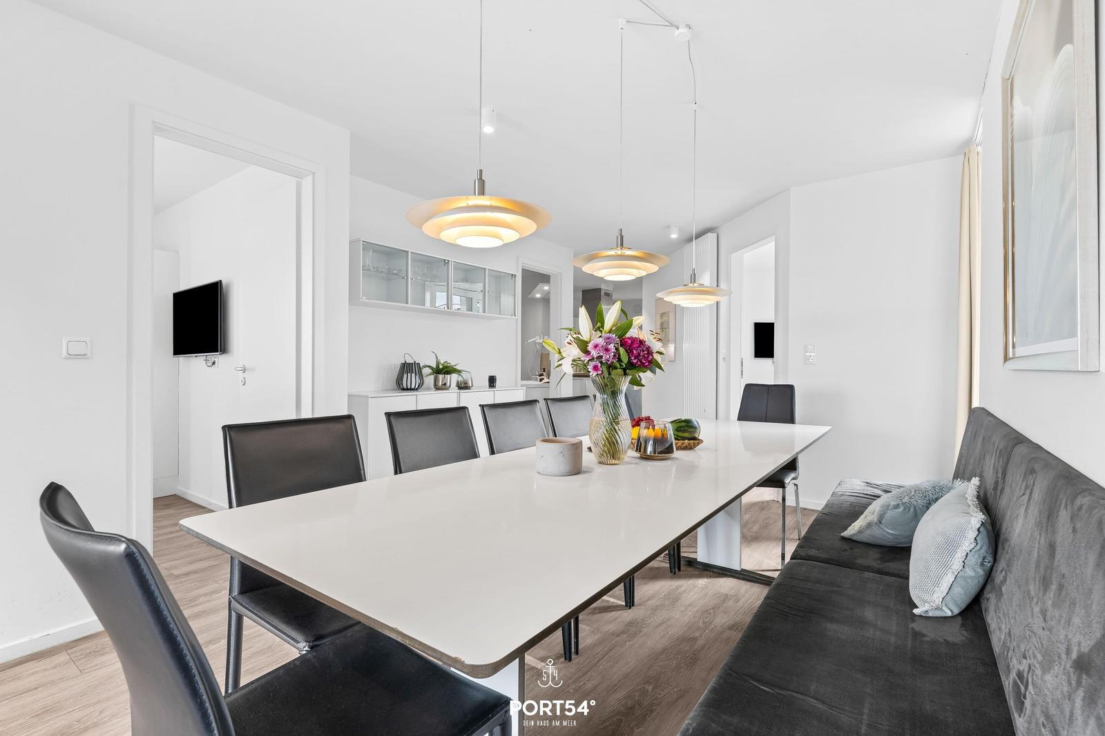 Essbereich mit langer Tischplatte, schwarzen Stühlen und grauem Sofa. Hängelampen und Blumen auf dem Tisch.