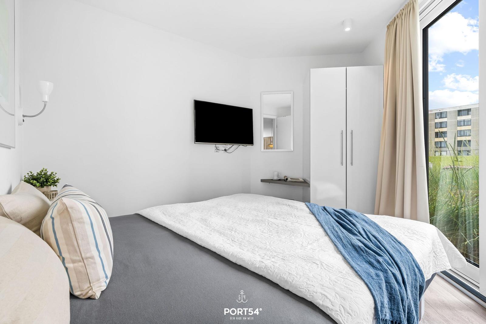 Schlafzimmer mit Bett, Fernseher und Garderobe. Großer Fensterblick auf Grünfläche.