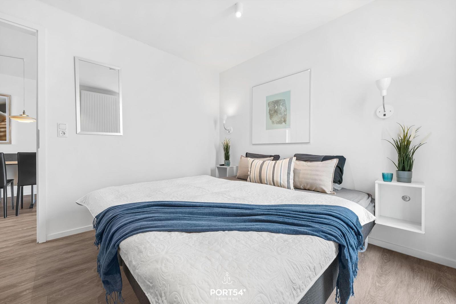 Schlafzimmer mit Bett, Wandlampen und Pflanzen. Holzboden und Wandspiegel sichtbar.
