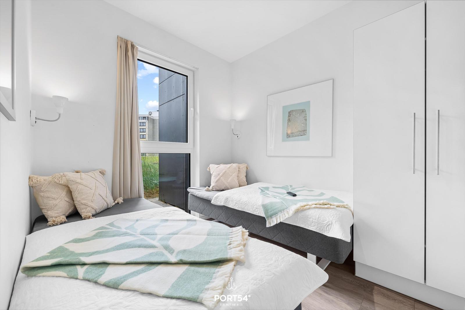 Zwei Betten mit Decken und Kissen. Fenster mit Blick. Weiße Wände und Kleiderschrank.