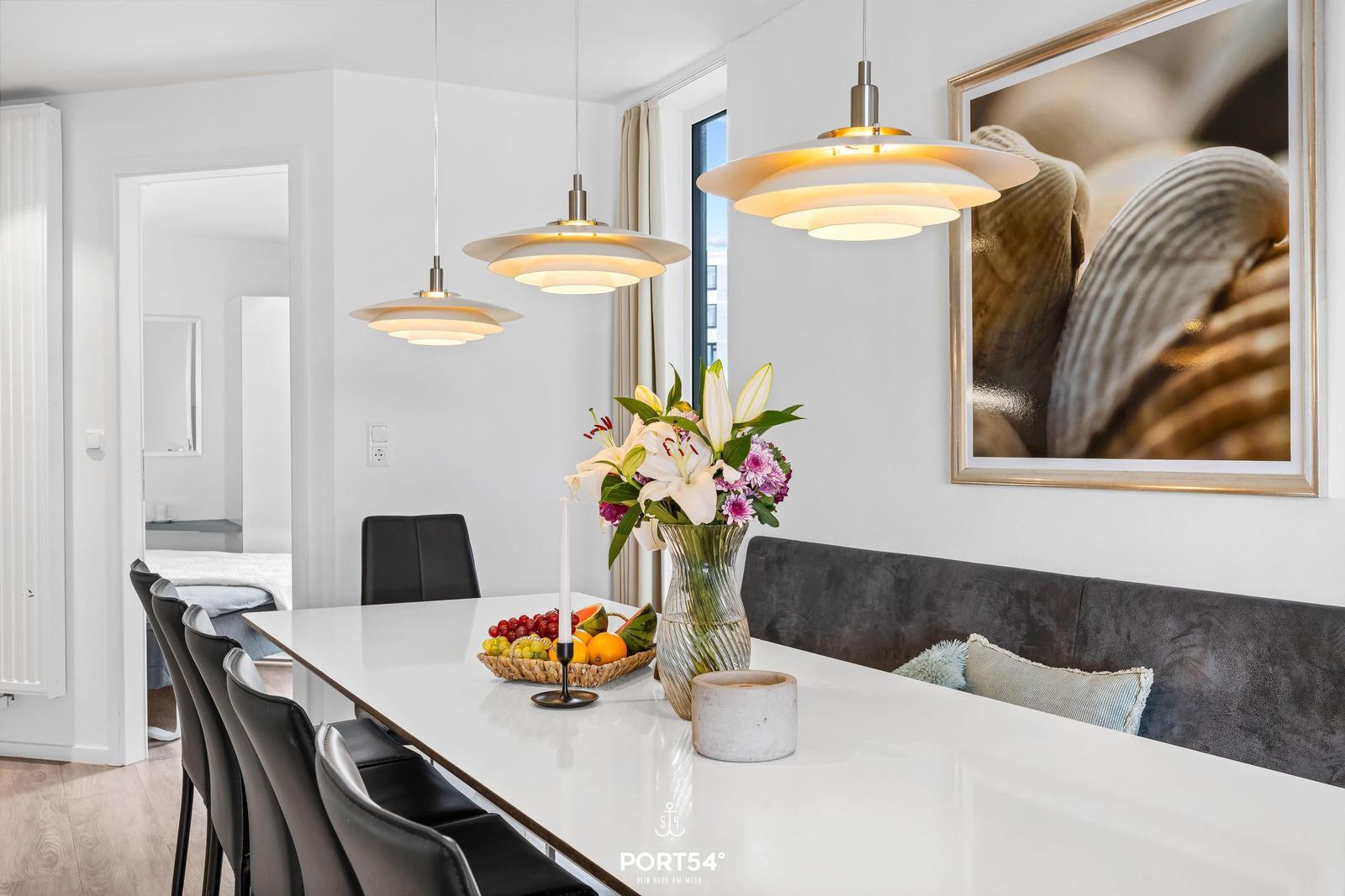Essbereich mit weißem Tisch, schwarzen Stühlen und modernen Lampen. Wandbild mit Muscheln.