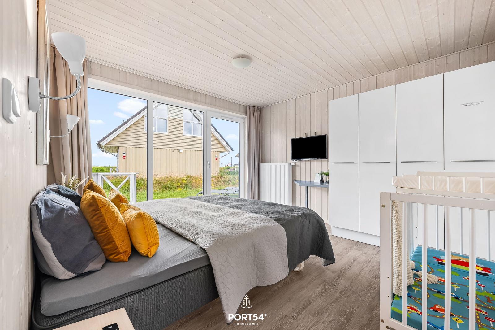 Schlafzimmer mit Bett, Kinderbett und Fernseher. Große Fenster mit Blick nach draußen.
