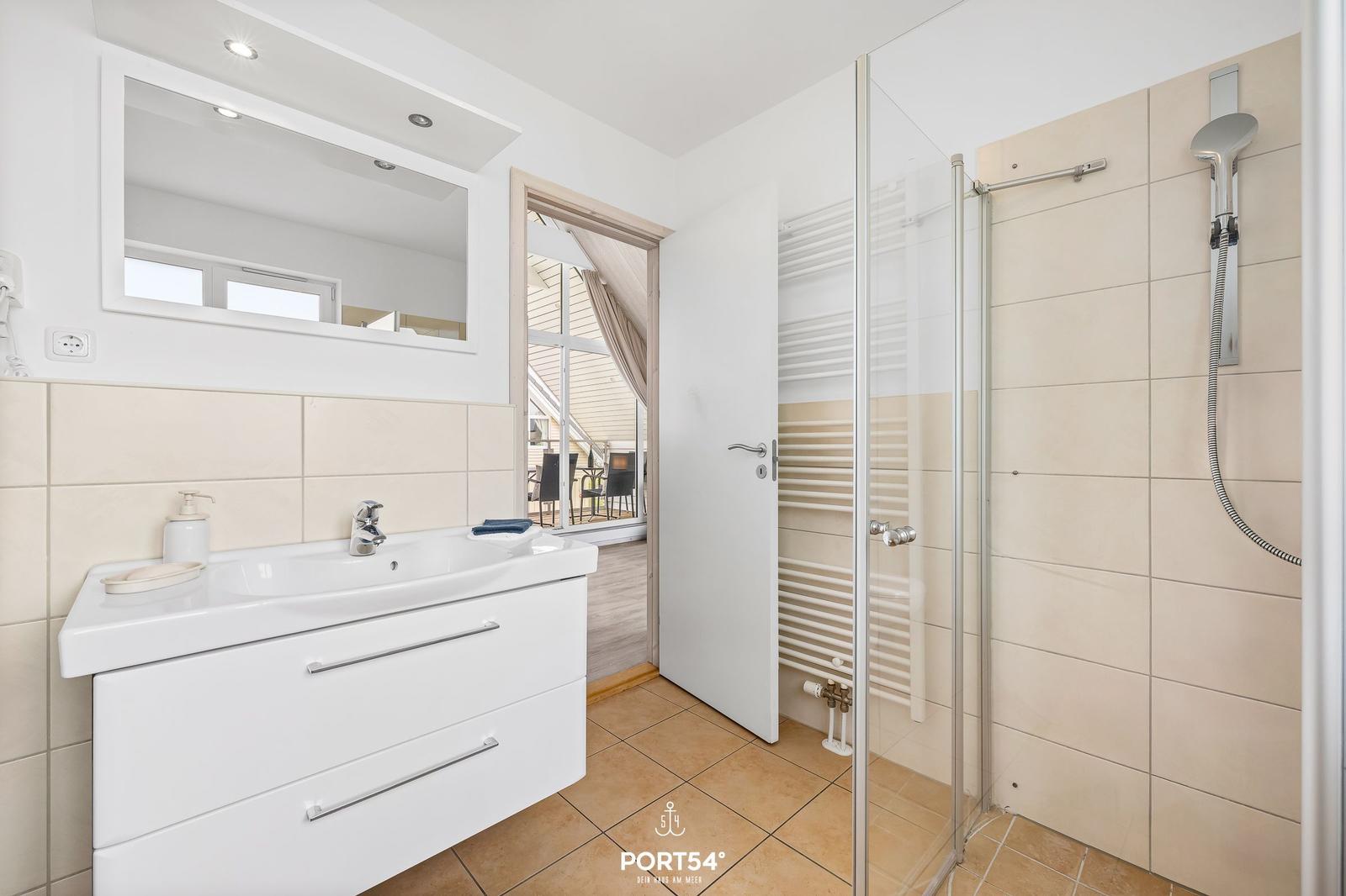 Badezimmer mit Dusche, Waschbecken und Spiegel. Tür führt zum Balkon.