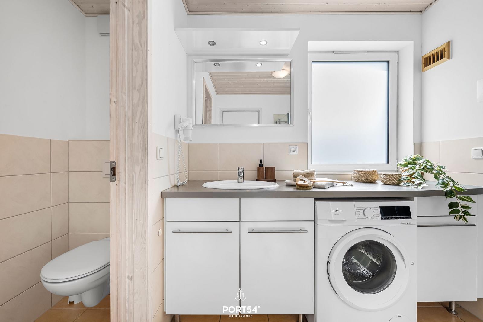 Badezimmer mit Waschmaschine, Spiegel und Toilette. Fenster mit Pflanze.