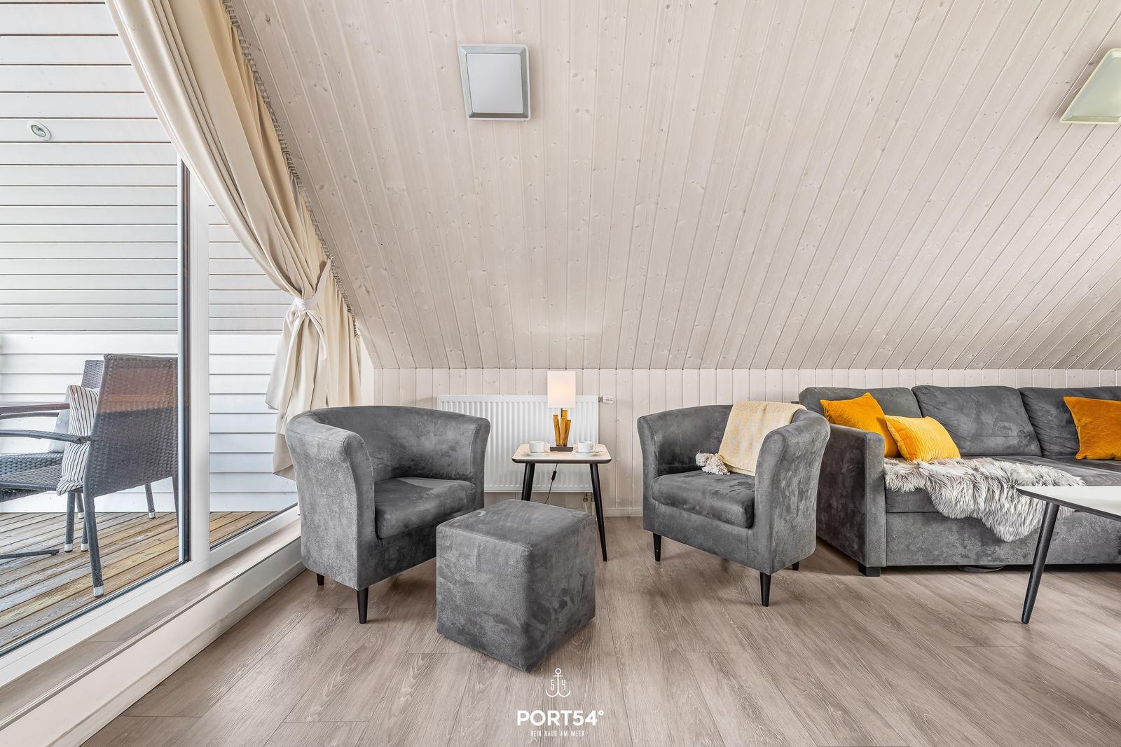 Wohnzimmer mit grauen Sesseln, Sofa und Holzboden. Große Fensterfront mit Blick auf Balkon.