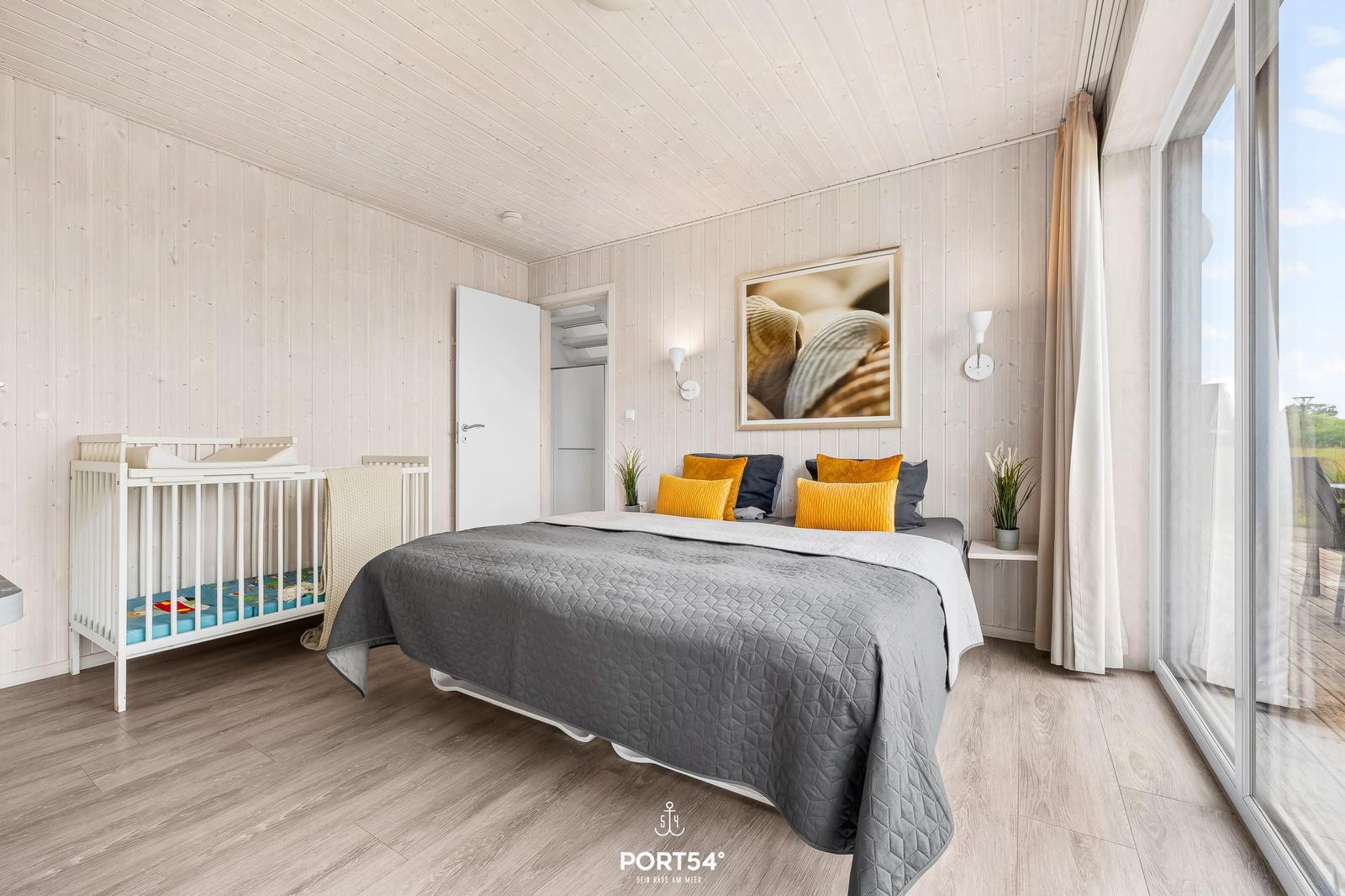 Schlafzimmer mit Bett, Wiege und großem Fenster. Holzwände und -boden.