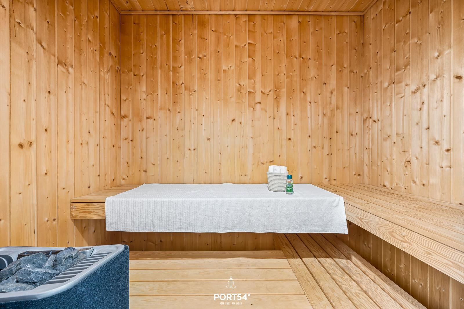Sauna mit Holzwand, Sitzbank und Deckenheizung. Auf der Bank liegen Handtücher und ein Spray.