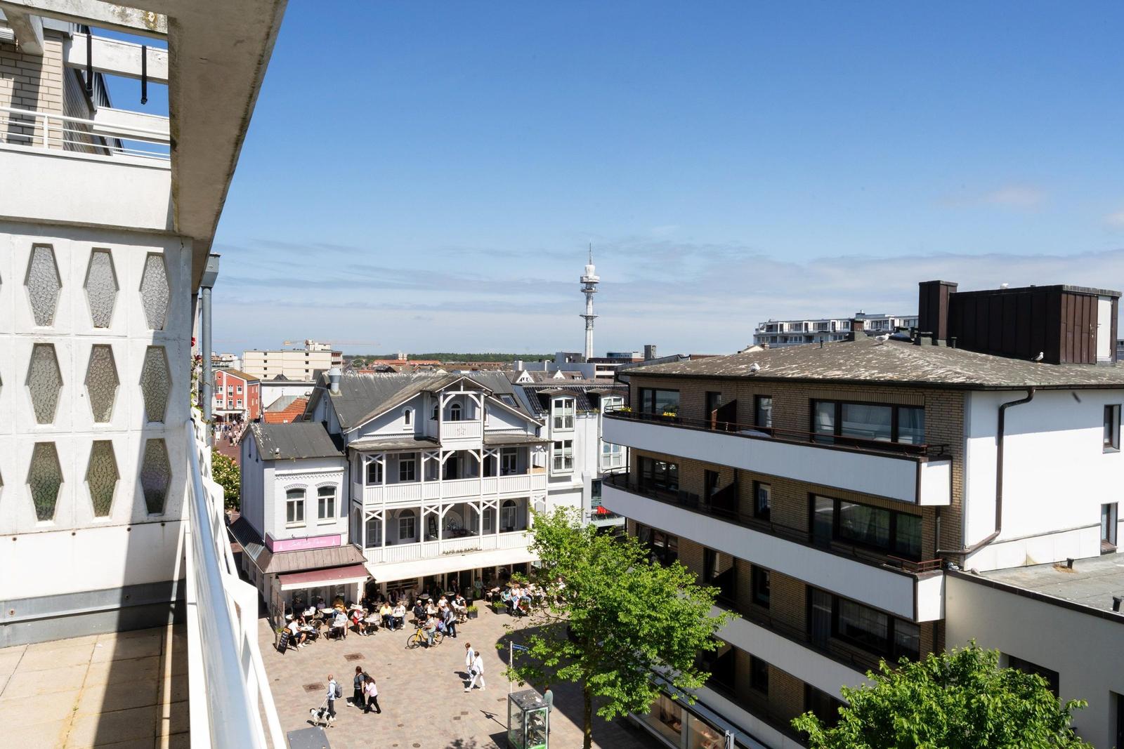 Aussicht auf belebten Platz mit Café, Bäumen und Stadtgebäuden unter blauem Himmel.