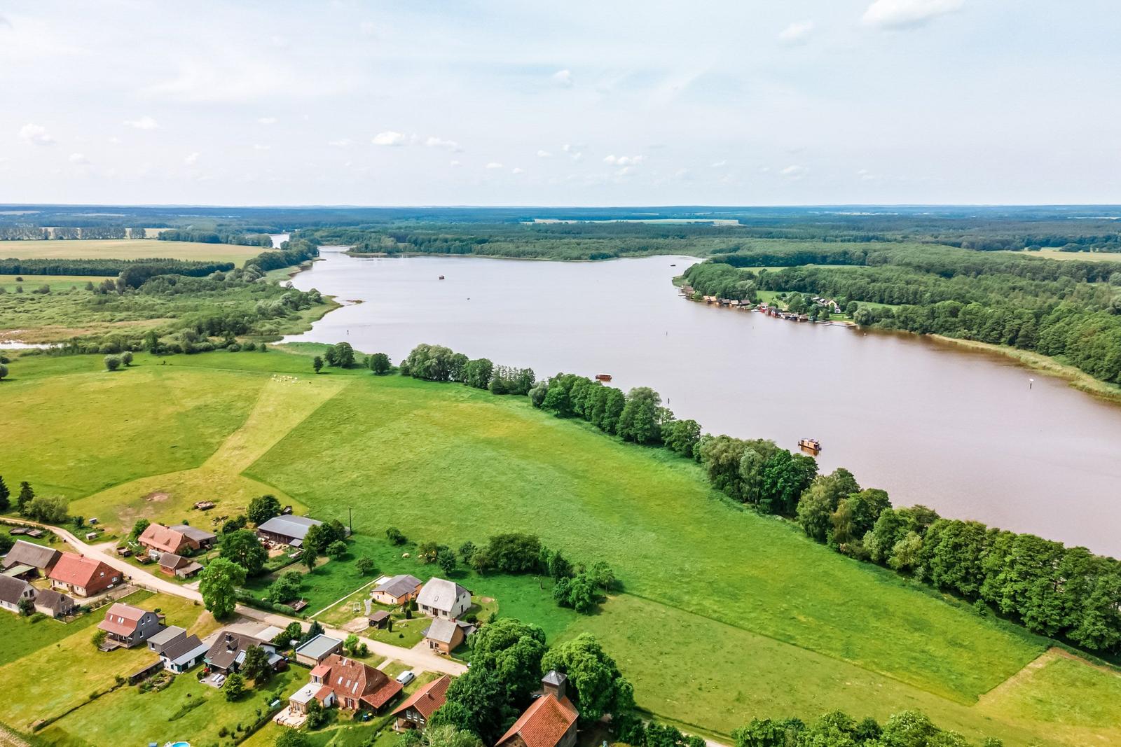 Ländliche Aussicht mit See, Wiesen und Bäumen. Einige Häuser sind am Ufer sichtbar.
