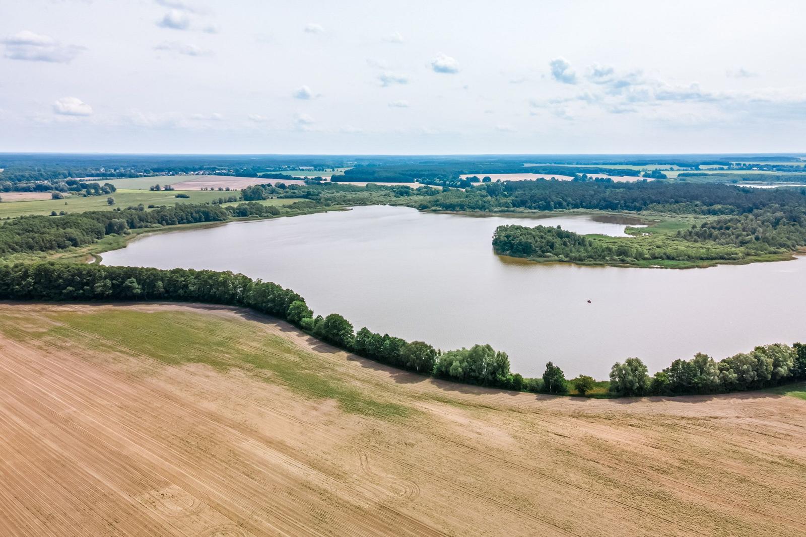 Aerial view of a lake surrounded by forests and agricultural fields under a partly cloudy sky.