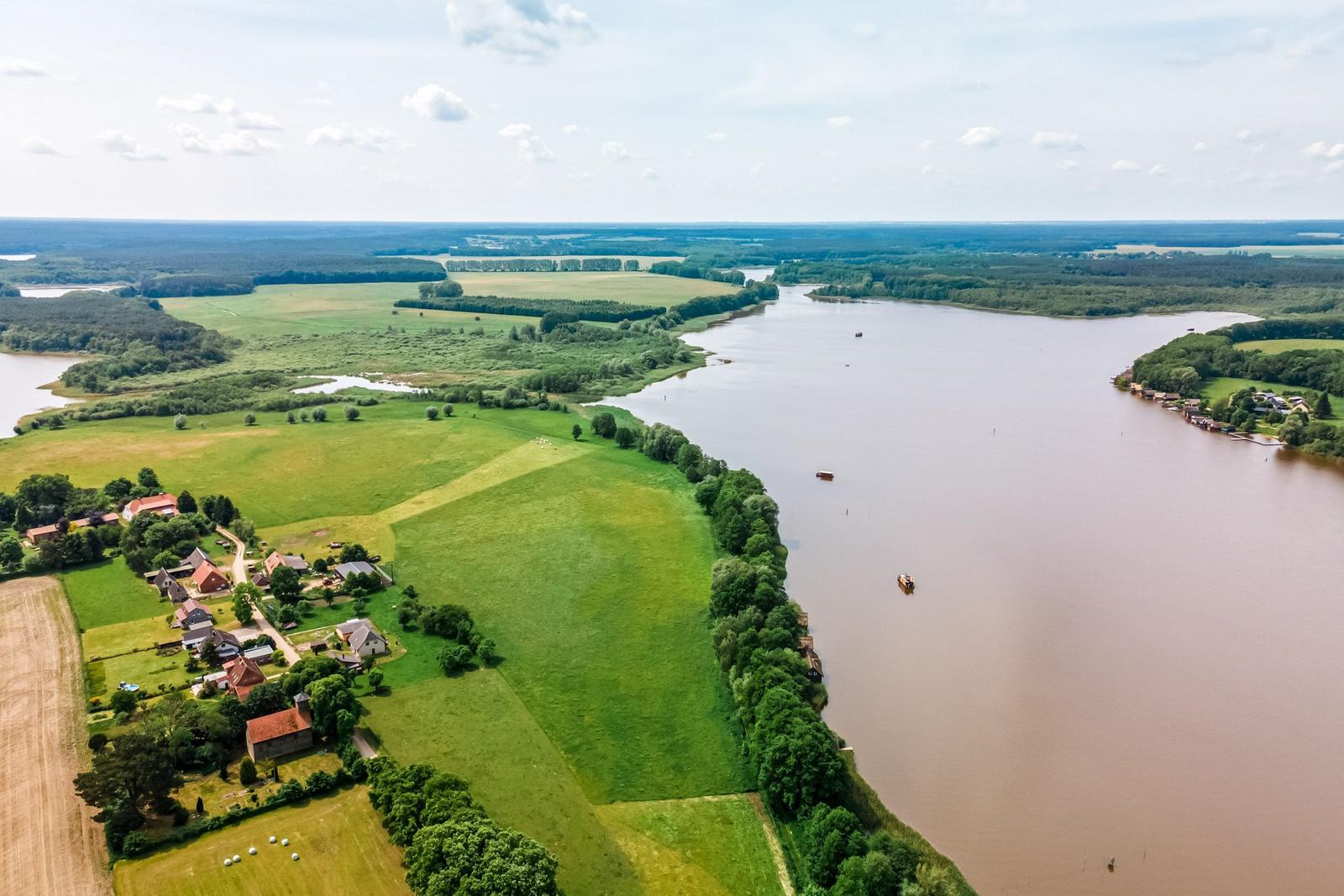 Aerielansicht eines Flusses mit grünen Feldern und einem Dorf an der Uferlinie.