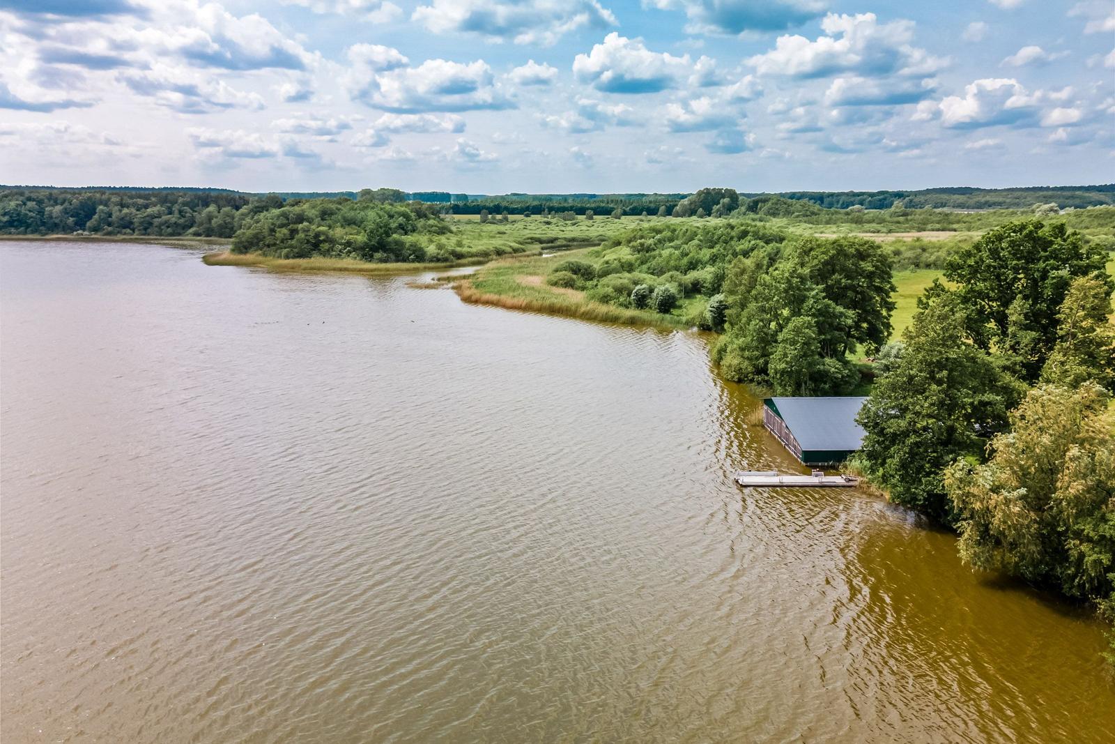 Seeufer mit kleinem Bootshaus und grüner Umgebung unter blauem Himmel.