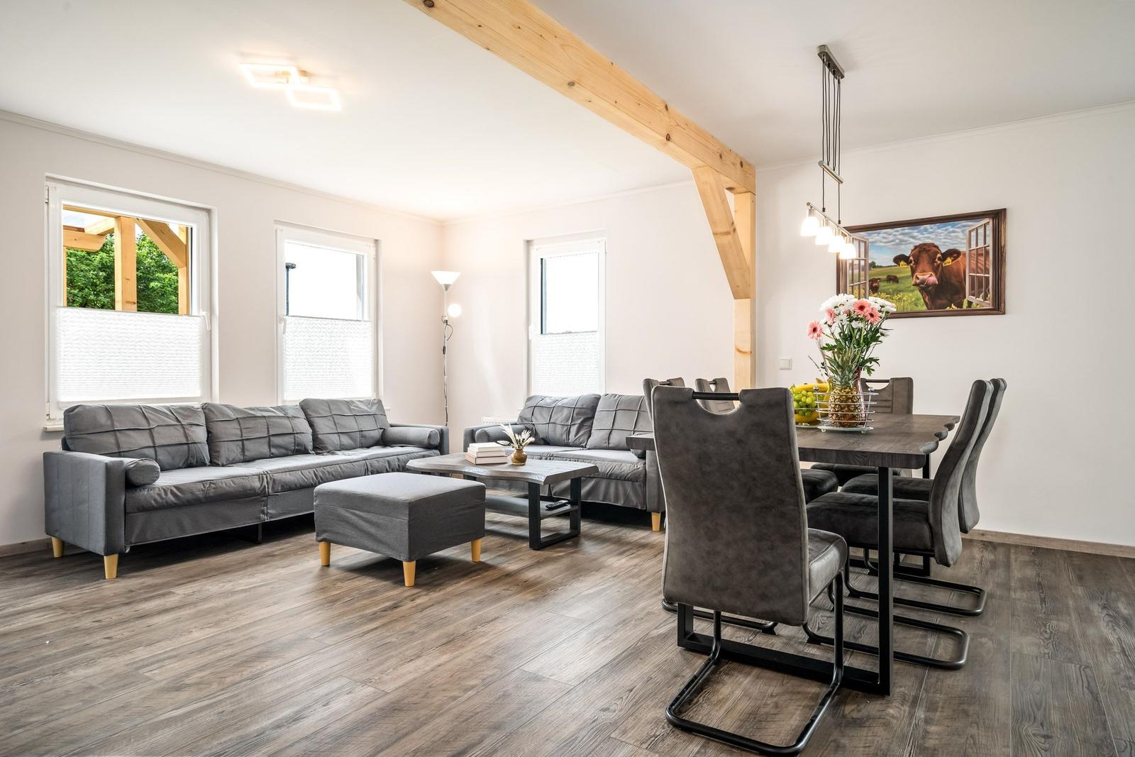 Wohn- und Essbereich mit grauen Sofas, Holzbalken und Tisch mit Stühlen.