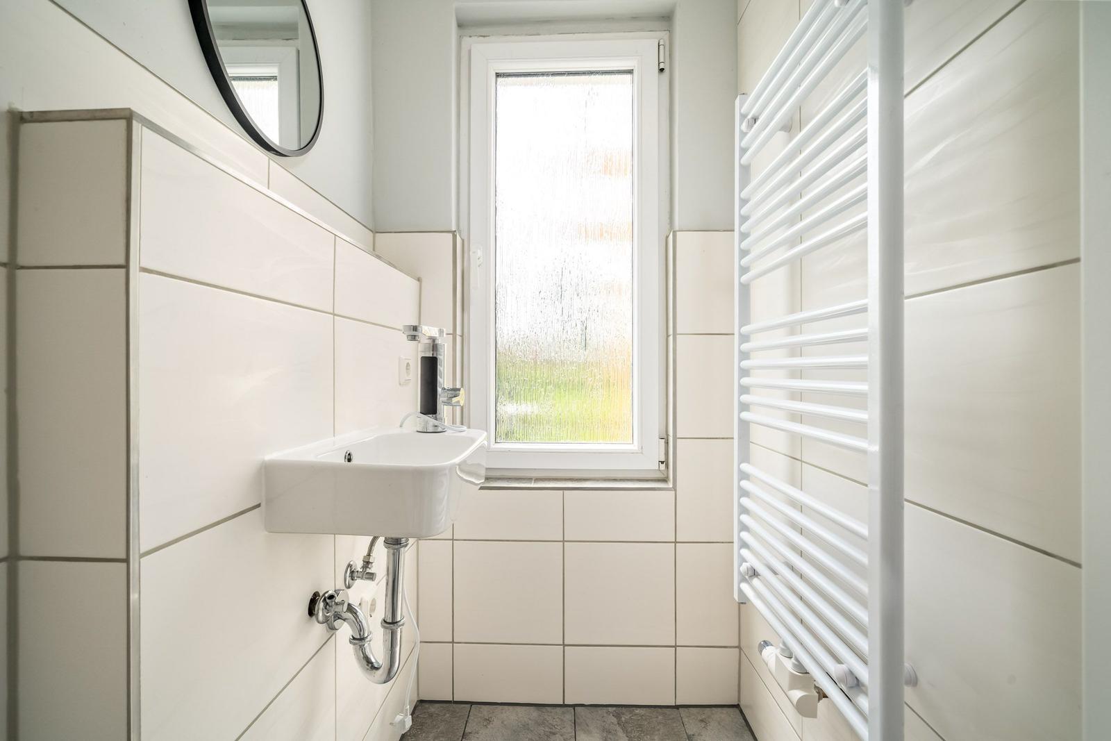 Badezimmer mit Waschbecken, Spiegel und Heizkörper. Fenster mit getöntem Glas.