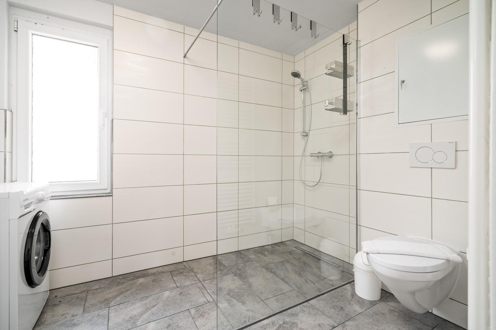 Badezimmer mit Dusche, Toilette und Waschmaschine. Weiße Fliesen und graue Bodenfliesen.