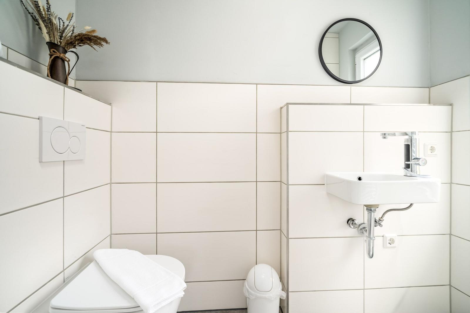Badezimmer mit Toilette, Waschbecken und Spiegel. Weiße Fliesen und moderne Einrichtung.