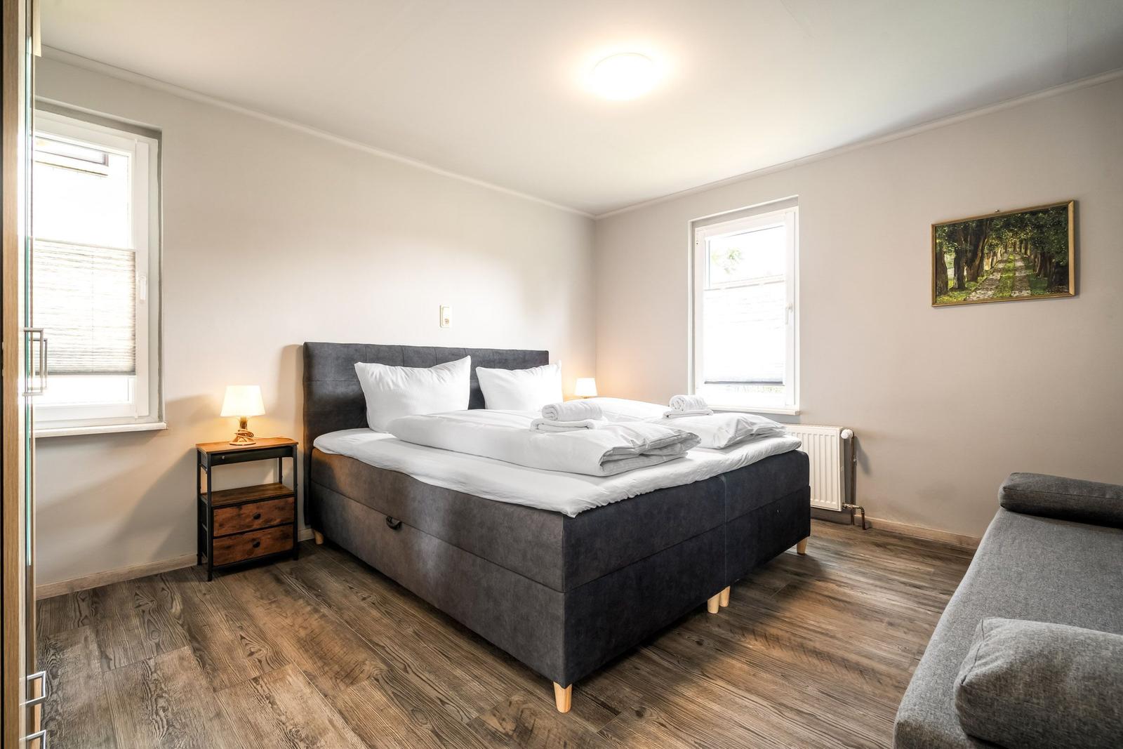 Schlafzimmer mit Bett, Nachttisch und Fenstern. Holzboden und Wandbild.