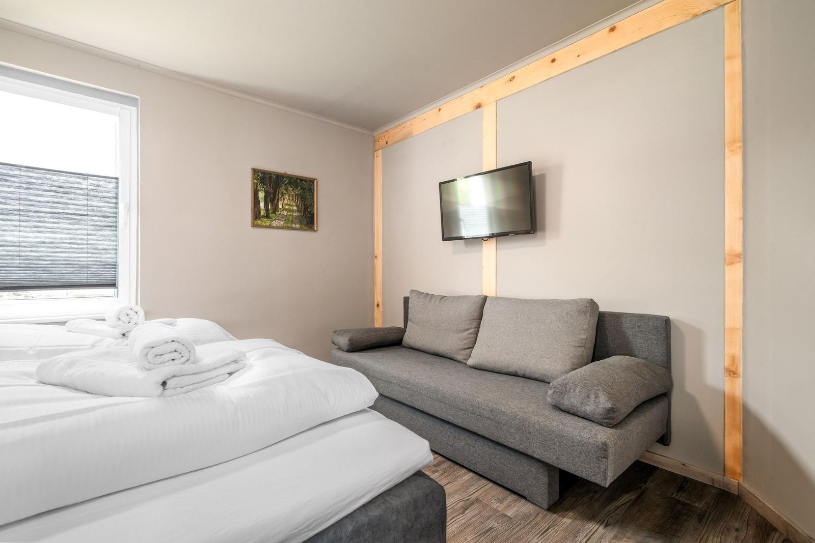 Schlafzimmer mit Bett, Sofa und Wandfernseher. Fenster mit Jalousie.