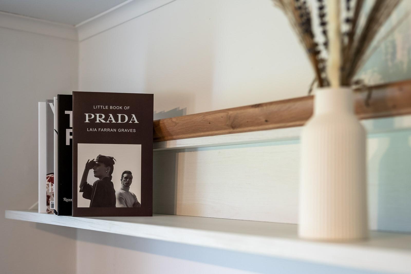 Ein Bücherregal mit einem Buch über Prada und einer weißen Vase mit Trockenpflanzen.